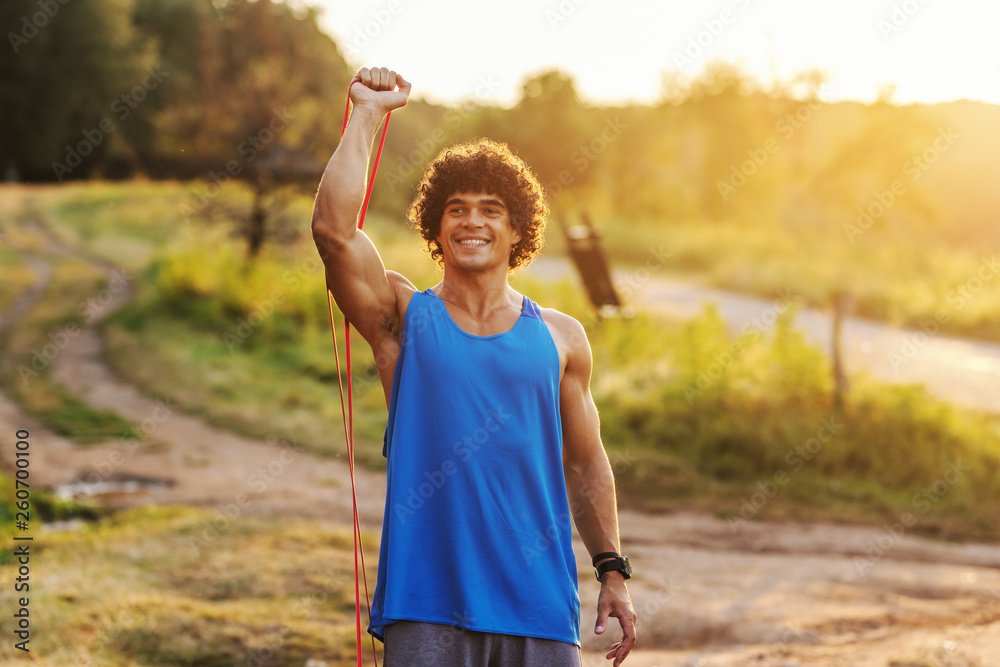 Smiling Caucasian sporty man with curly hair doing stretching exercises with rope in nature in sunny summer day.