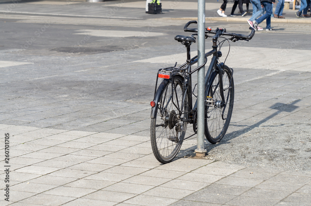 a black bicycle tethered to a sign, in the background you can see the legs of people a black bicycle tethered to a sign, in the background you can see the legs of people