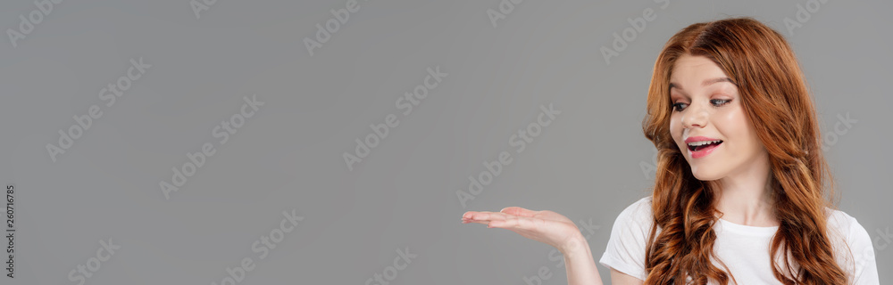 panoramic shot of beautiful redhead girl gesturing with hand and presenting something isolated on grey with copy space panoramic shot of beautiful redhead girl gesturing with hand and presenting something isolated on grey with copy space