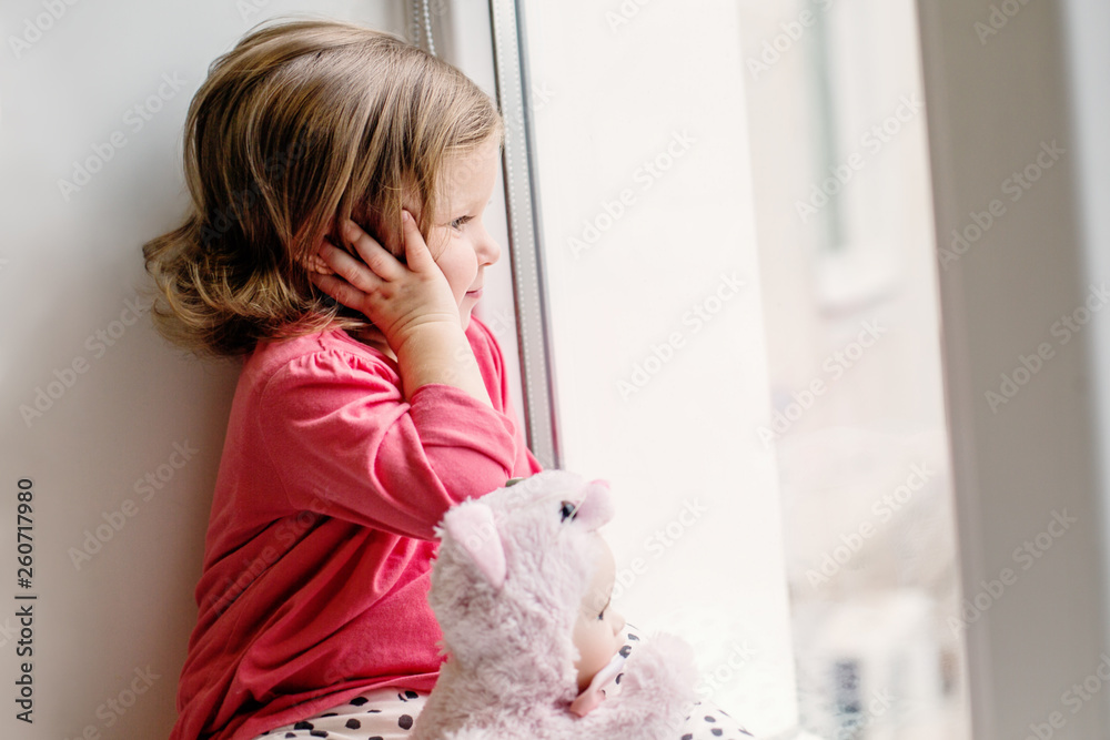 little baby girl sitting on a window sill looks out the window along with a doll