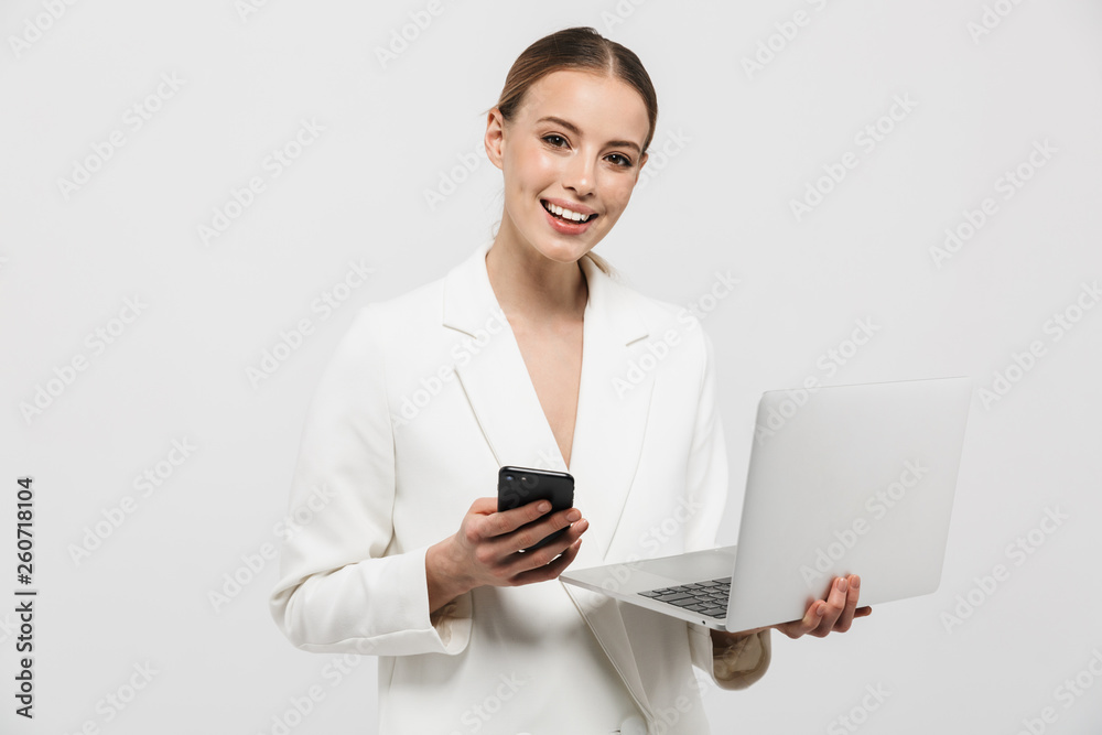 Photo of pretty businesswoman 20s wearing elegant jacket smiling while holding laptop and smartphone Photo of pretty businesswoman 20s wearing elegant jacket smiling while holding laptop and smartphone