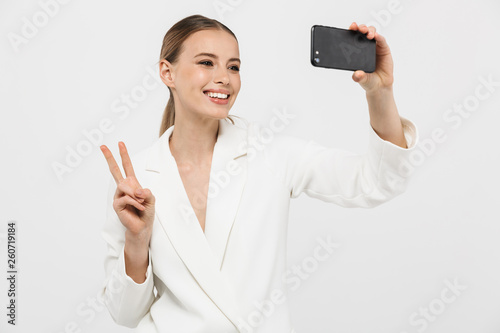 Photo of pleased businesswoman 20s wearing elegant jacket holding cell phone and taking selfie photo