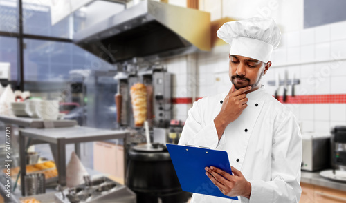 cooking, profession and people concept - male indian chef in toque reading menu on clipboard and thinking over kebab shop kitchen background