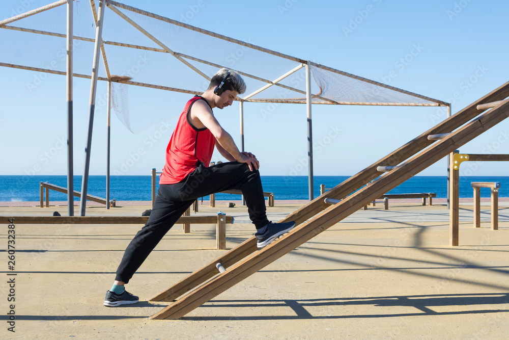 Man stretching his muscles before starting to practice outdoor sports, with urban gym machines made of wood, near the sea.