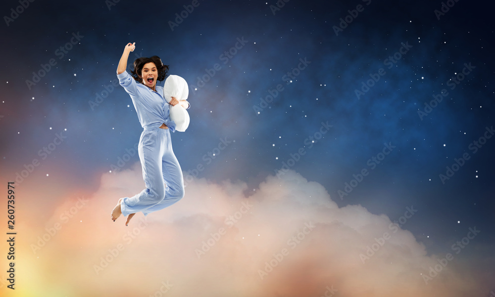 fun, people and bedtime concept - happy young woman full of energy in blue pajama holding pillow and jumping over starry night sky background
