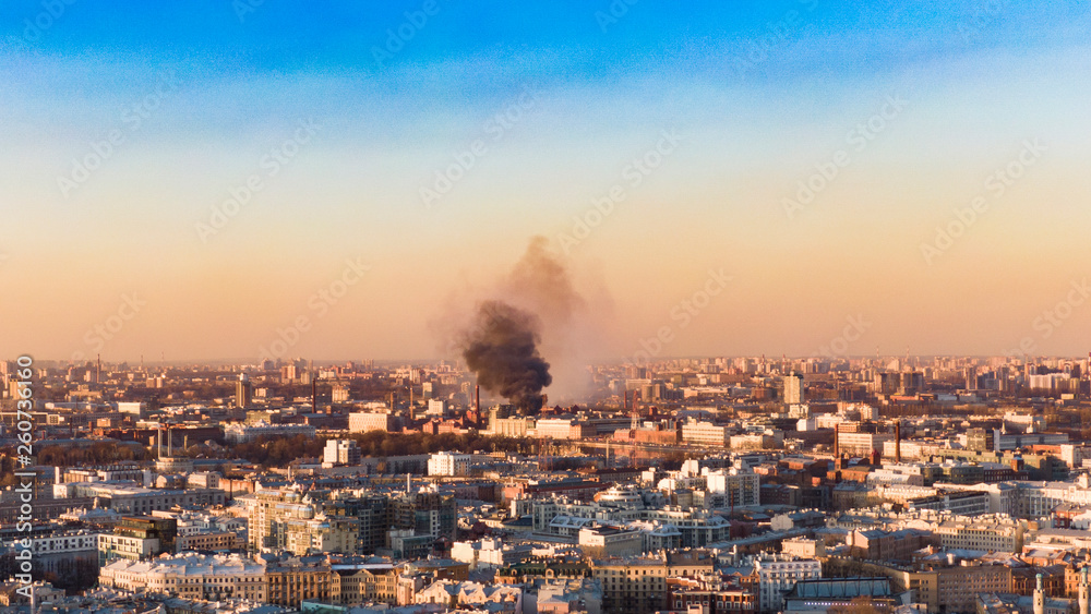 Fire in the city, a column of black smoke rises above horizon, aerial view