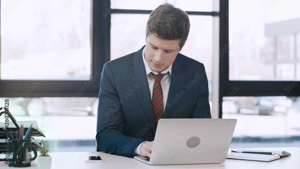 handsome man typing on laptop, looking at smartphone, talking, smiling and writing in notebook in modern office handsome man typing on laptop, looking at smartphone, talking, smiling and writing in notebook in modern office