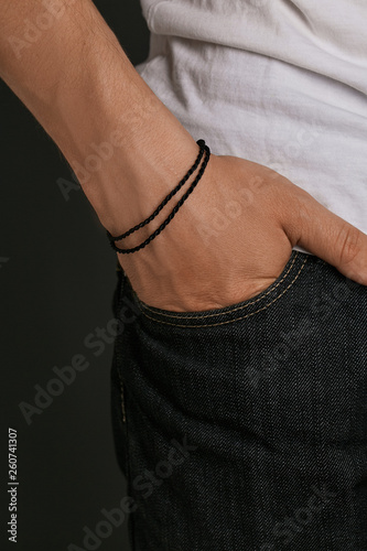 Cropped closeup shot of man's hand, wearing black lucky rope bracelet with 2 rows of thick string. The guy is wearing jeans and shirt, putting his hand into the pocket, posing on dark background.