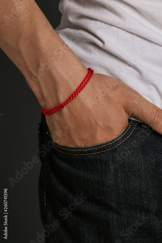 Cropped half-turn shot of man's hand with tanned skin, wearing red lucky rope bracelet. The guy is wearing black jeans and white shirt, putting his hand into the pocket, posing on dark background.