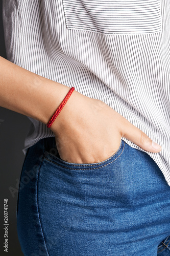 Cropped closeup shot of woman's hand, wearing red lucky rope bracelet with fishtail plaiting. The lady is wearing jeans and stripy shirt, putting her hand into pocket, posing against dark background.