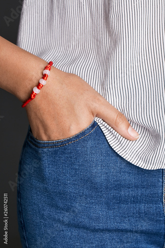 Cropped closeup shot of lady's hand, wearing red lucky rope bracelet, adorned with 3 milky beads. The girl is wearing jeans and stripy shirt, putting hand into pocket, posing on dark background.