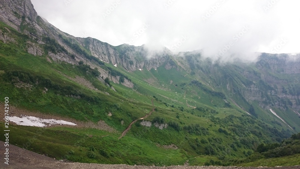 Panorama of the mountains and of the Aibga ridge with low clouds. Remains of snow and fresh green grass on the mountains near the ski resort Rosa Khutor in Krasnaya Polyana. Sochi, Russia.