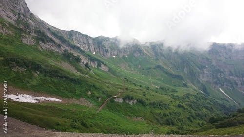 Panorama of the mountains and of the Aibga ridge with low clouds. Remains of snow and fresh green grass on the mountains near the ski resort Rosa Khutor in Krasnaya Polyana. Sochi, Russia.
