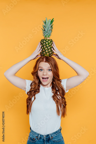 cheerful redhead girl looking at camera and posing with pineapple on head isolated on yellow