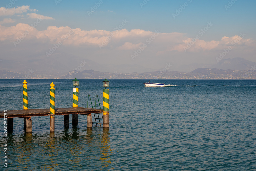 Scenic view of Lake Garda with a wooden pier with vintage striped poles, a passing motorboat and a cloudy blue sky, Sirmione, Lombardy, Italy