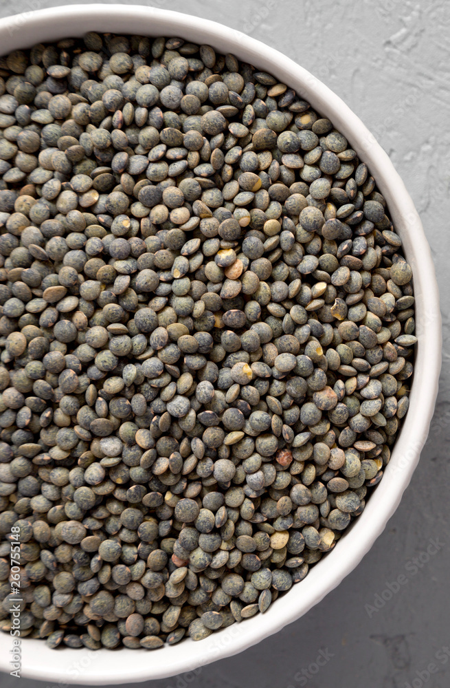 Full bowl of dry green french lentils on a gray surface, top view. Overhead, from above, flat lay. Full bowl of dry green french lentils on a gray surface, top view. Overhead, from above, flat lay.