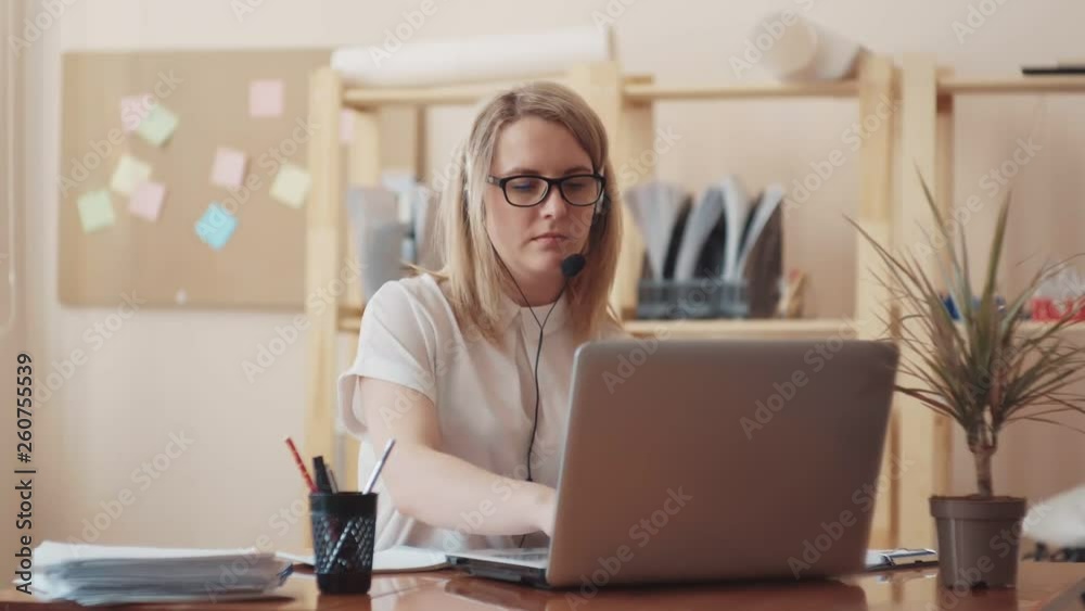 sweet girl of Caucasian appearance with glasses and a white blouse works for the laptop and the headset is communicating with the client, writing with a pen his words sweet girl of Caucasian appearance with glasses and a white blouse works for the laptop and the headset is communicating with the client, writing with a pen his words