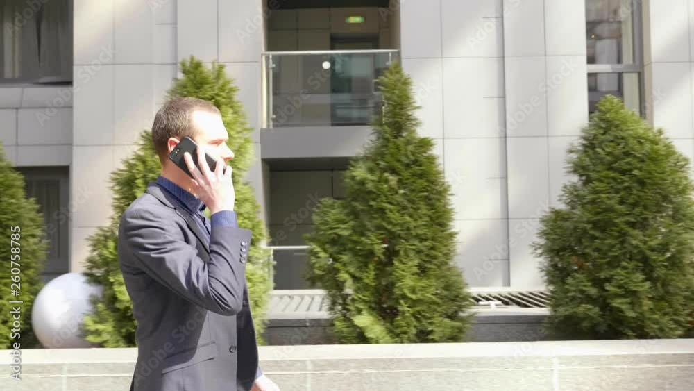 A young businessman walking down the street and talks over the phone call. A guy in a classic suit leads a conversation on the smartphone. Small green trees on the background. 4K footage. A young businessman walking down the street and talks over the phone call. A guy in a classic suit leads a conversation on the smartphone. Small green trees on the background. 4K footage.