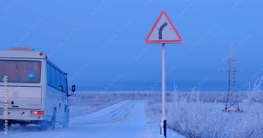 The white bus is moving along a snowy country road that goes to the right, as the road sign says The white bus is moving along a snowy country road that goes to the right, as the road sign says