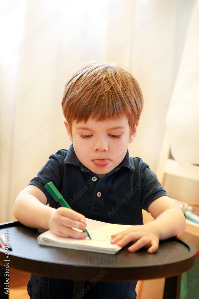 Child is drawing and painting with felt pen on paper of spiral notebook on small wooden table in living room at home. Childhood. Cute little boy and kid draws pencil and colorful markers in preschool. Child is drawing and painting with felt pen on paper of spiral notebook on small wooden table in living room at home. Childhood. Cute little boy and kid draws pencil and colorful markers in preschool.