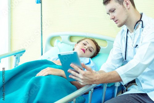 Young friendly caucasian pediatrician male doctor is examining patient woman with tablet pc on bed in hospital  consultation with stethoscope in clinic office. Healthcare and medical concept