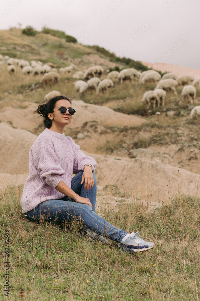 Happy young woman in a sweater against herds of sheep in the mountains
