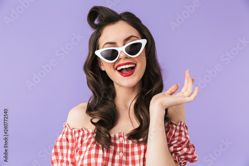 Portrait of joyful pin-up woman 20s in american style wear and retro sunglasses smiling at camera