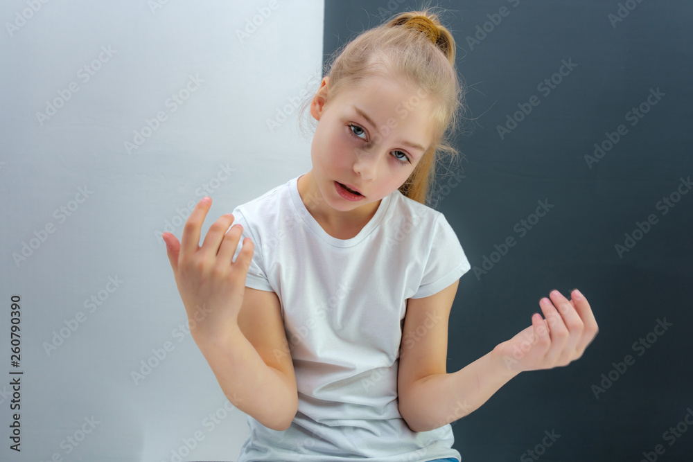 A concept portrait of a cute pretty blonde teen girl with long hair is sitting in a white T-shirt against a gray-white background and talking. A concept portrait of a cute pretty blonde teen girl with long hair is sitting in a white T-shirt against a gray-white background and talking.