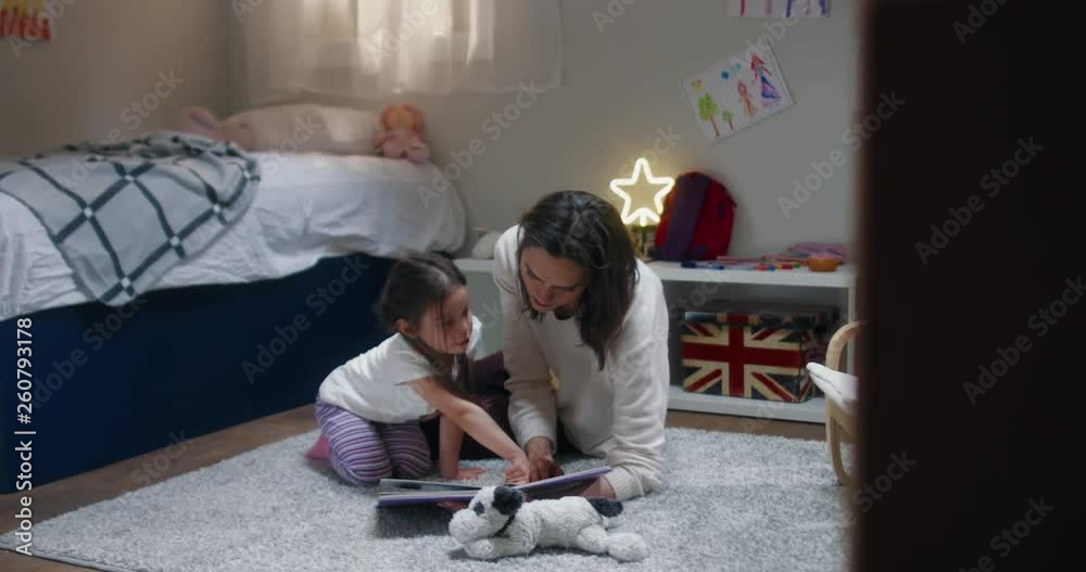 DOLLY Portrait of cute little girl reading a book together with her mother at home, evening shot, bedroom interior. 4K UHD RAW FOOTAGE DOLLY Portrait of cute little girl reading a book together with her mother at home, evening shot, bedroom interior. 4K UHD RAW FOOTAGE