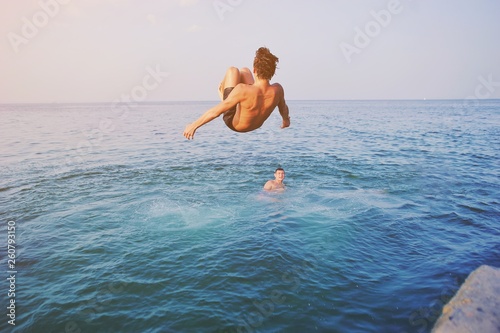 Young man jumping off cliff into blue water ocean at sunset. Active outdoor, holiday adventure, tourism action, healthy summer joy, Fun activity lifestyle. Crazy adult guy in swimwear fly from climb