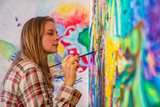 Joyful young female artist painting on the wall, using brush and bright acrylic paints