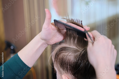 Barber shop. Hairdresser makes hairstyle to a boy with hair scissors and black comb. 