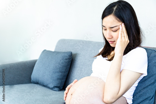 Pregnant Asian woman with hand on forehead suffering headache sitting on a couch in the living room.