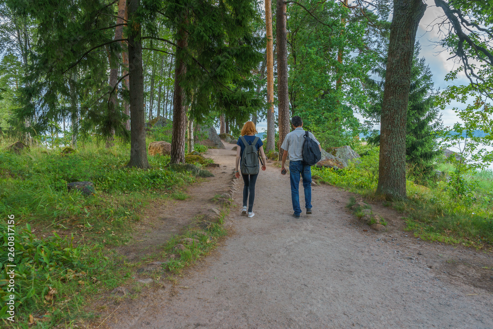 Dark-haired middle-aged man and red-haired young lady walk along forest road. Tourists on the beautiful landscape background. Monrepos Park, Vyborg, Russia. Travel concept Dark-haired middle-aged man and red-haired young lady walk along forest road. Tourists on the beautiful landscape background. Monrepos Park, Vyborg, Russia. Travel concept