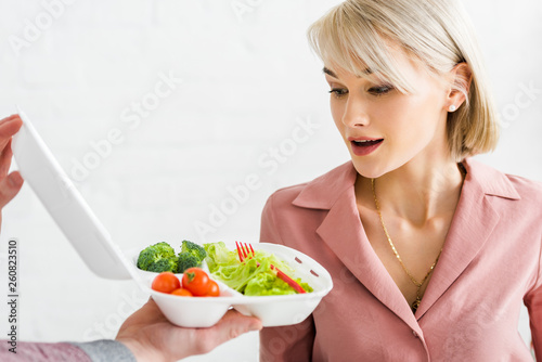 cropped view of man holding takeaway box with vegetables near surprised young woman