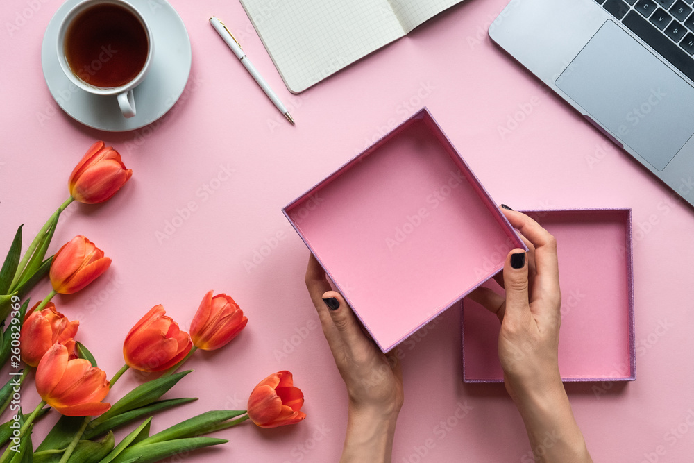 Obraz premium Female hands holding open empty box on pink background. Background with cup of tea, laptop and flowers