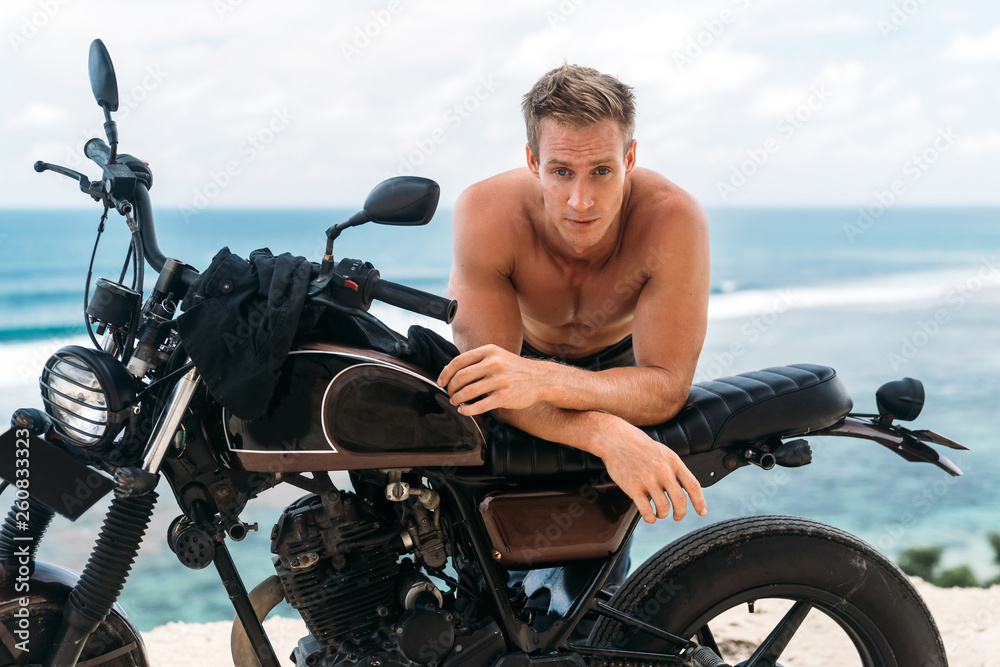 Portrait of sexy athletic man with naked torso on custom motorbike with ocean at background