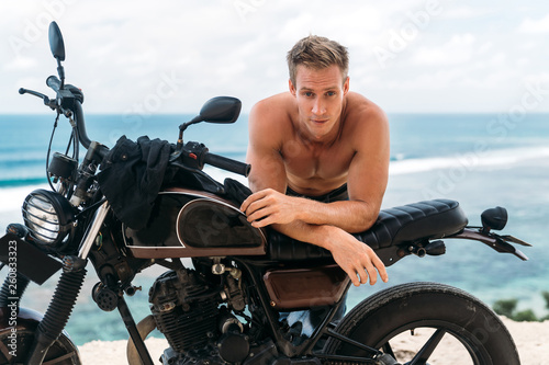 Portrait of sexy athletic man with naked torso on custom motorbike with ocean at background © romannoru