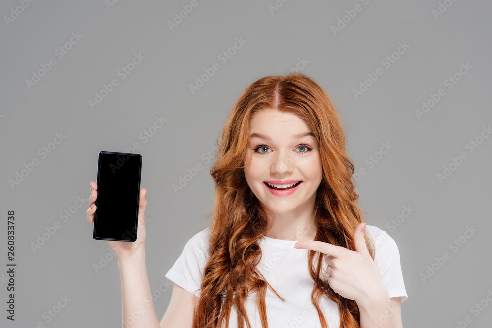 Naklejka premium beautiful redhead girl pointing with finger at smartphone with blank screen, looking at camera and smiling isolated on grey