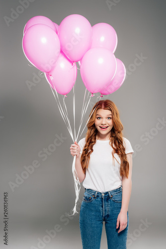 beautiful surprised girl with red hair holding pink air balloons and looking at camera on grey