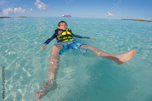 Young boy floating in life jacket in crystal clear blue water with cruise ship in background.