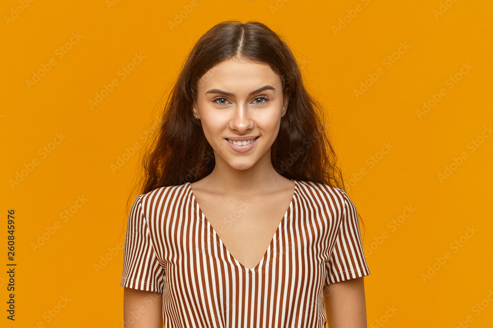 Summer, youth and happiness concept. Adorable joyful girl with broad happy smile and loose long hair enjoying leisure time, posing against blank yellow wall background wearing nice striped dress