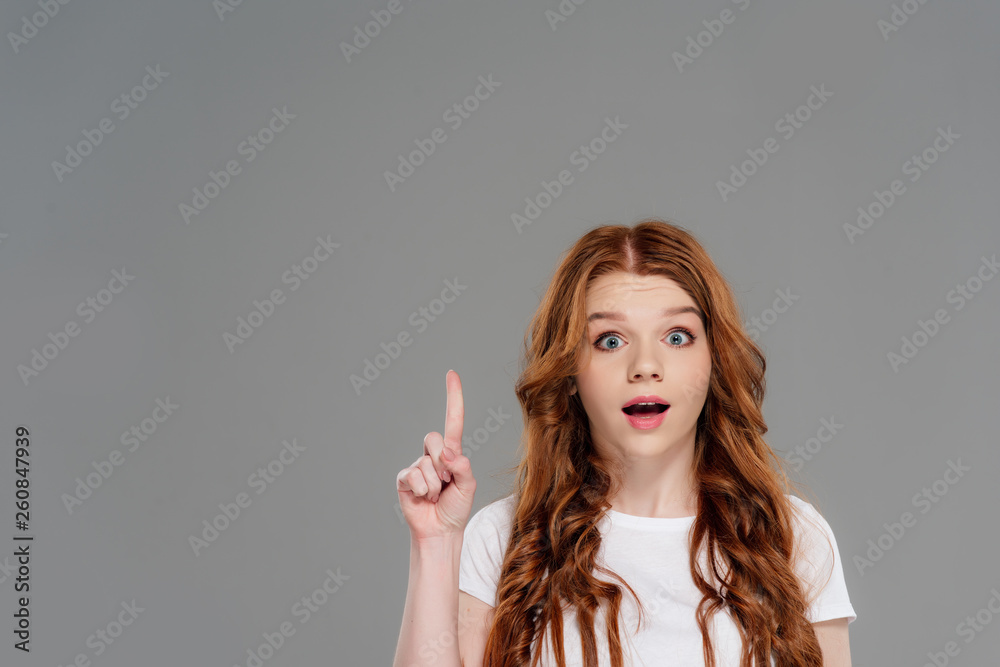 beautiful surprised redhead girl looking at camera and showing idea gesture isolated on grey with copy space beautiful surprised redhead girl looking at camera and showing idea gesture isolated on grey with copy space