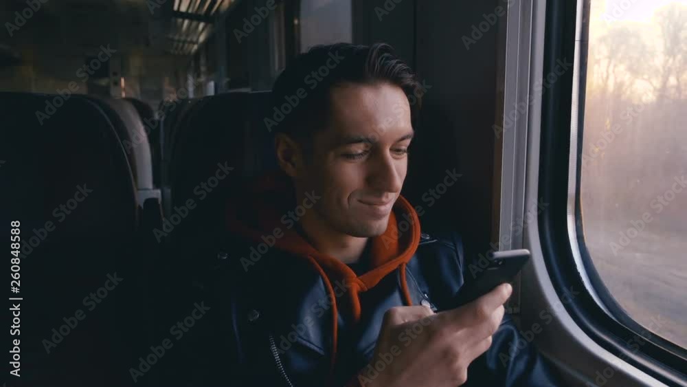 Young man using smartphone during train ride and sitting opposite the window with beautiful sunset