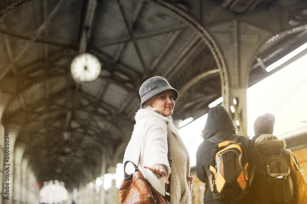 A beautiful woman in a hat on a train station background and a platform with a clock looks back and shows to follow her, holding out her hand. Go past the passengers of the train.