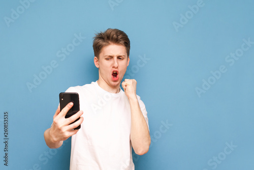 Portrait of a joyful man with a smartphone in his hands, emotionally reacts, looks at the screen, wears a white T-shirt and stands on a blue background. Copyspace