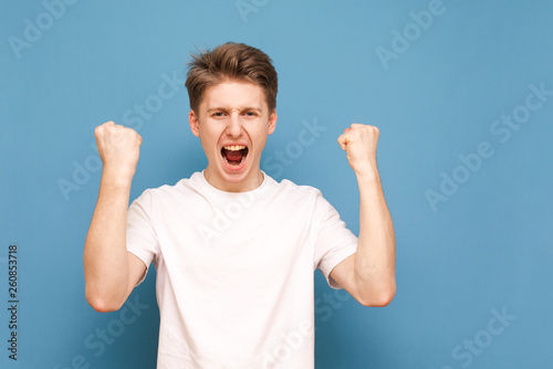 Expresive young man in a white T-shirt rejoices on a blue background, lifting his arms and shouting with joy. Boy emotionally rejoices, hands raised, isolated on a blue background. Copyspace