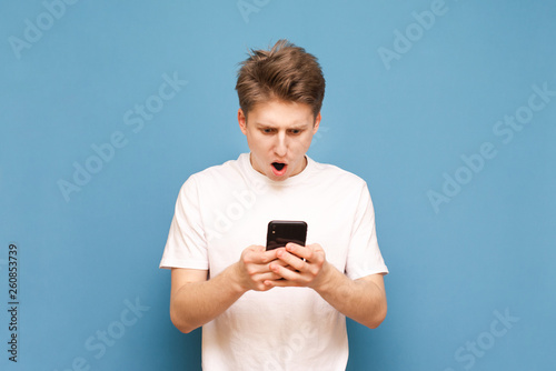 Amazed young man in a white T-shirt uses a smartphone and reacts emotionally to the blue background. Shocked guy is surprised to look at the screen of a smartphone. Isolated
