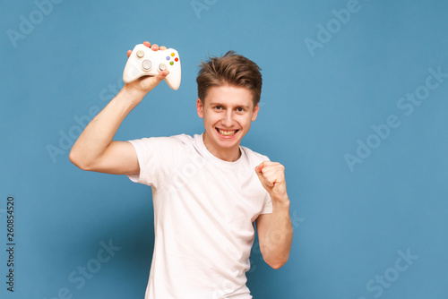 Portrait of a happy gamer plays video games, rejoices in victory on a blue background, looks into the camera and rejoicing. Joyful gamer with a joystick in his hand is isolated on a blue background.