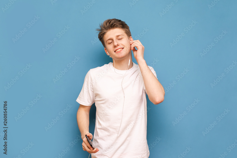 Happy young man wearing a white T-shirt enjoys music in his headphones with his eyes closed, holding a smartphone in his hands, isolated on a blue background. Copyspace Happy young man wearing a white T-shirt enjoys music in his headphones with his eyes closed, holding a smartphone in his hands, isolated on a blue background. Copyspace